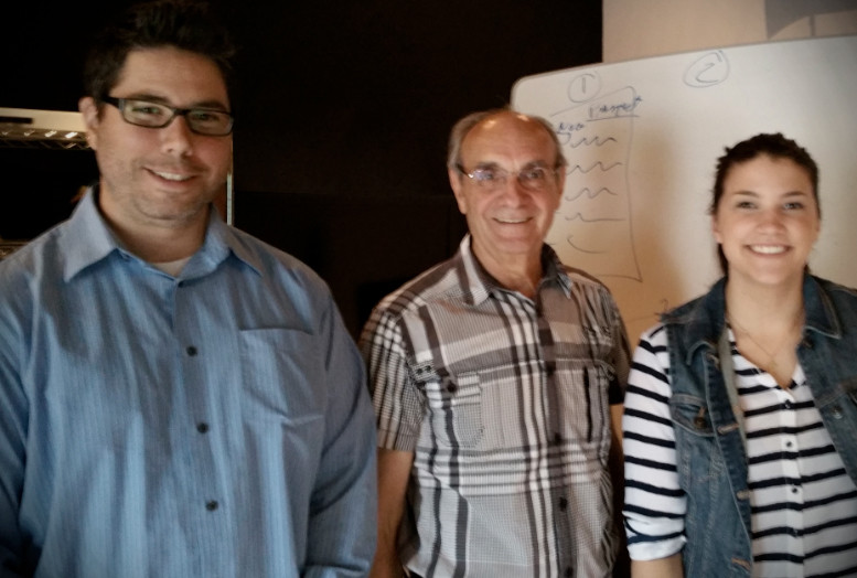 Trois personnes souriantes devant un tableau blanc avec des notes manuscrites dans une salle sombre.