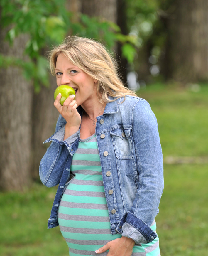 Femme enceinte en veste en jean mangeant une pomme verte dans un parc verdoyant.