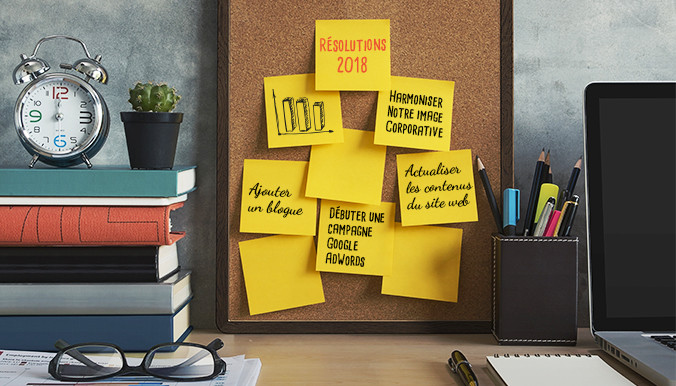 Table de bureau avec horloge, livres, lunettes, ordinateur portable et tableau avec notes de résolutions 2018.