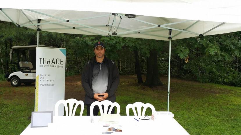Homme debout sous un kiosque blanc avec une bannière Thrace et une table avec brochures en plein air.
