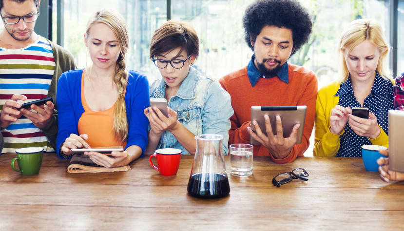 Groupe diversifié de cinq personnes utilisant des appareils mobiles assises à une table en bois avec tasses et carafe.