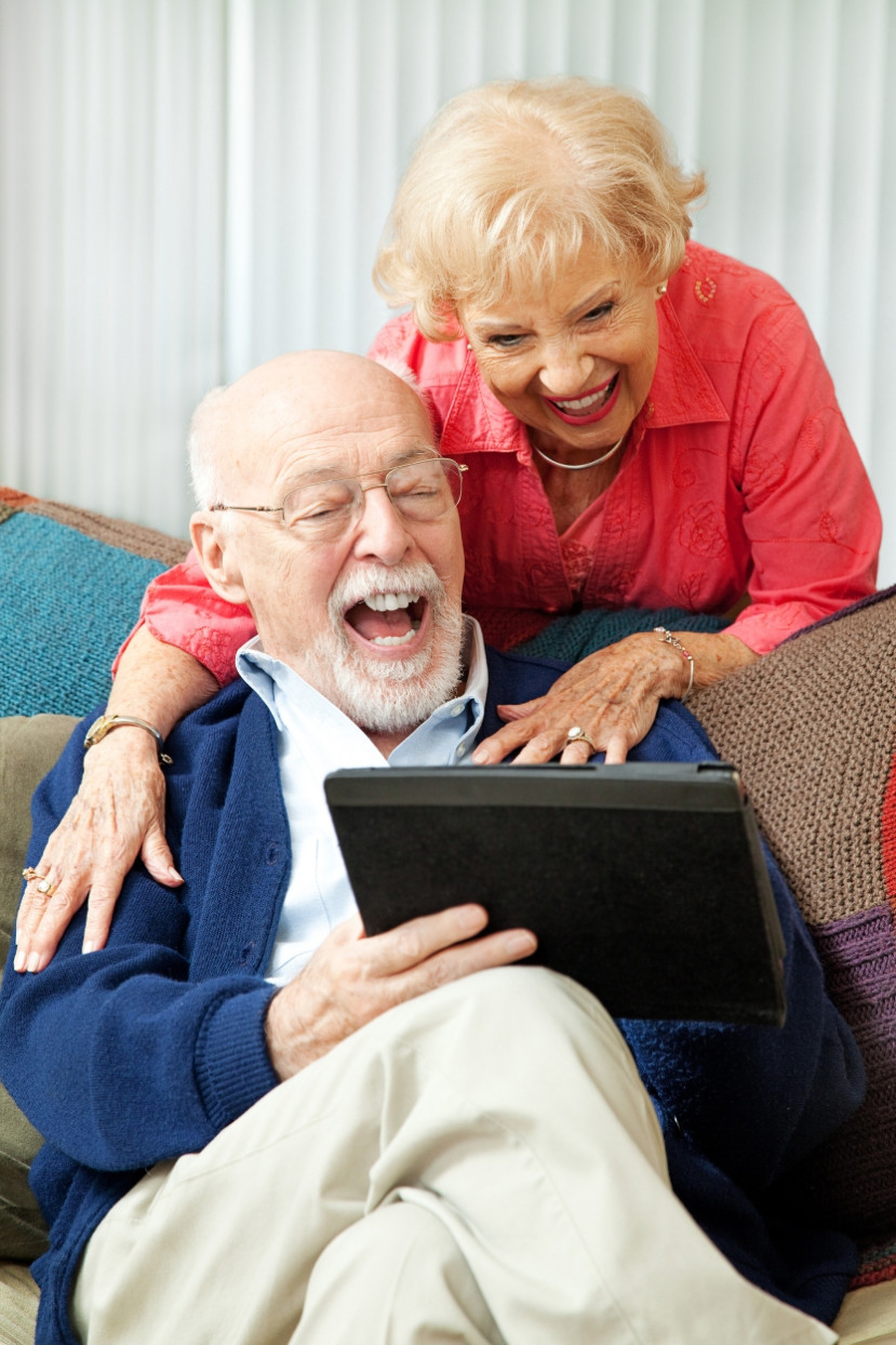 Couple aîné souriant utilisant une tablette numérique assis sur un canapé avec coussins colorés.