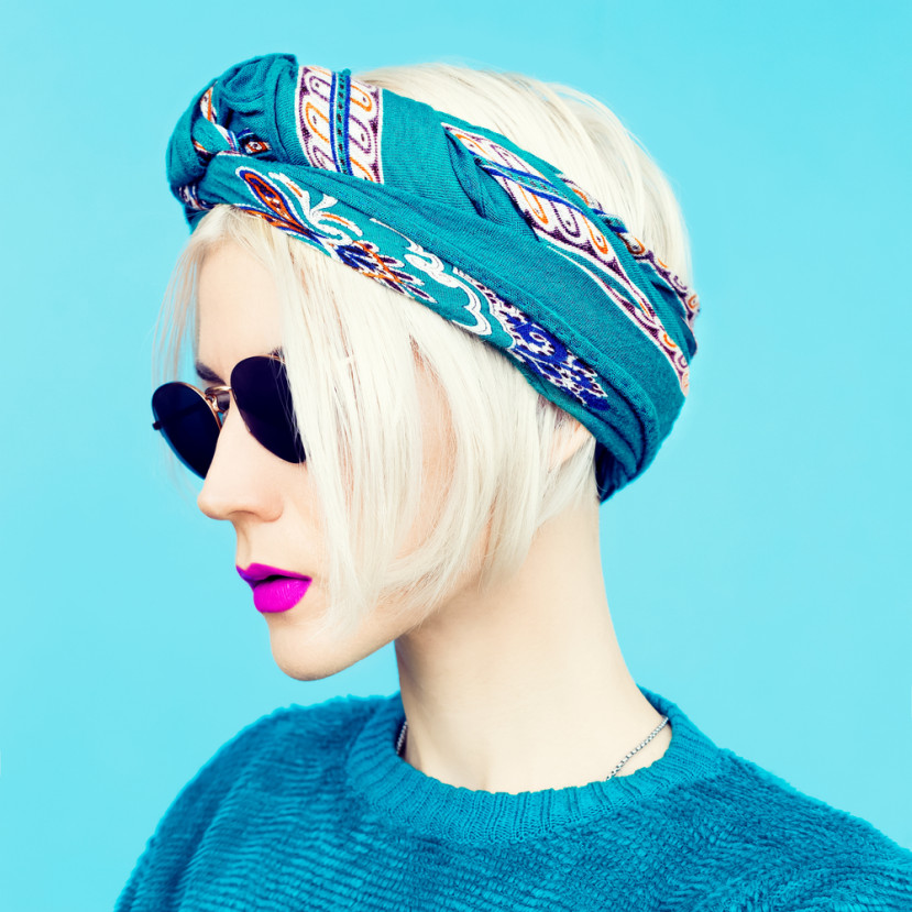 Femme aux cheveux blonds courts portant un bandeau turquoise, lunettes noires et rouge à lèvres violet vif.