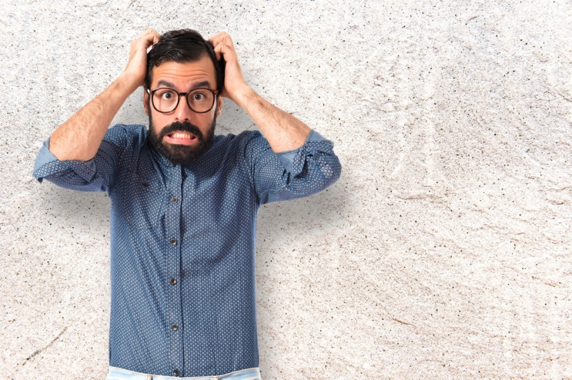 Homme avec lunettes et chemise bleue tenant sa tête avec expression de stress devant un mur clair.