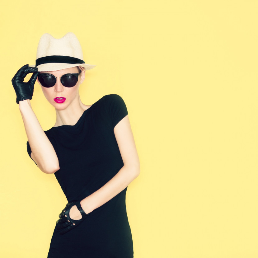 Femme élégante en robe noire, chapeau blanc, lunettes de soleil et gants noirs sur fond jaune.